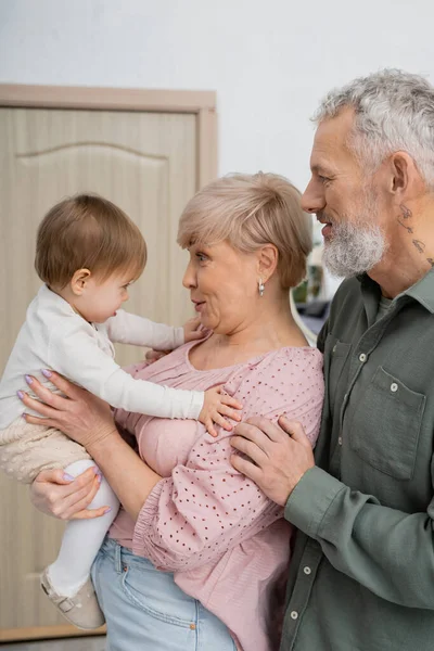 Seitenansicht einer glücklichen Frau, die ihre kleine Enkelin in der Nähe des bärtigen Ehemannes zu Hause betrachtet — Stockfoto