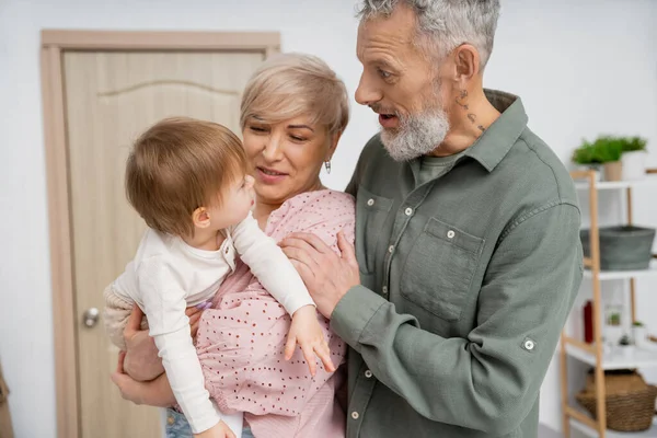 Erstaunt bärtiger Mann schaut Kleinkind Enkelin in den Händen von Frau in Flur der Wohnung — Stockfoto