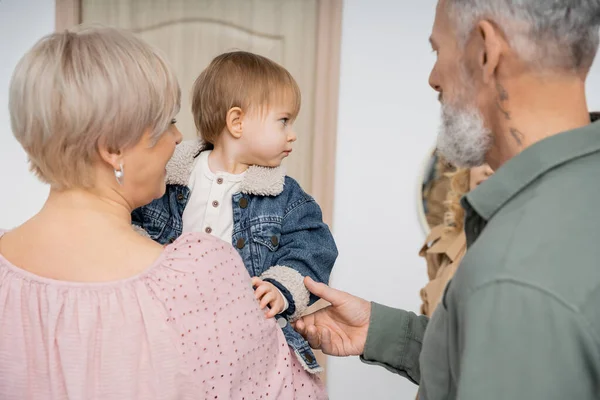 Hombre barbudo tocando la mano de la pequeña nieta en chaqueta de mezclilla cerca de la familia y puerta de entrada del apartamento — Stock Photo