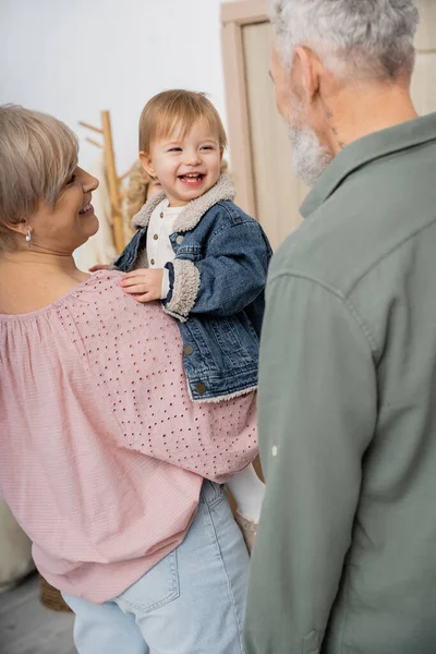 Middle aged woman holding cheerful granddaughter in denim jacket near family at home — Stock Photo