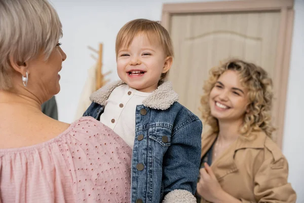 Радісна дитина в джинсовій куртці посміхається в руках бабусі біля щасливої матері і вхідних дверей на розмитому фоні — стокове фото