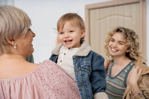 Middle aged woman holding happy little girl in denim jacket near blurred daughter smiling at entrance door — Stock Photo