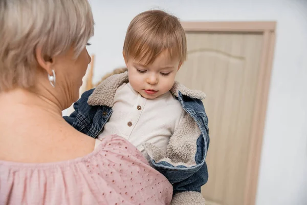 Middle aged woman holding little granddaughter in denim jacket in hall of apartment — Stock Photo