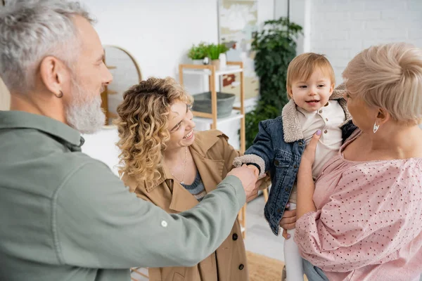 Bearded man holding hand of toddler granddaughter near happy family in living room — Stock Photo
