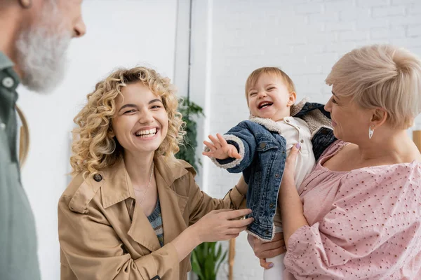 Überglückliche Frau im Trenchcoat lächelt in die Kamera neben kleiner Tochter und glücklichen Eltern zu Hause — Stockfoto