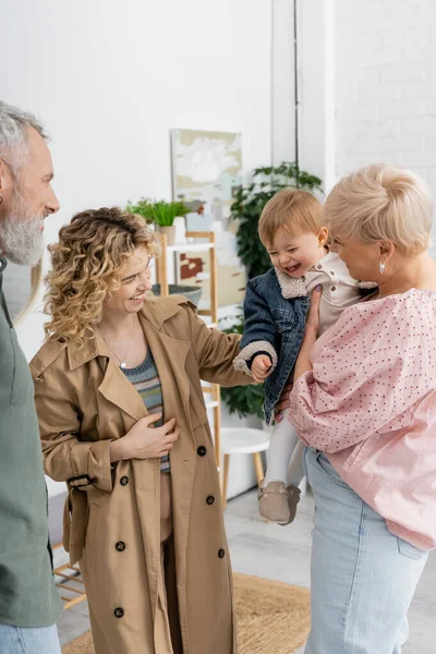Задоволена жінка в траншеї пальто посміхається біля щасливих батьків і маленької дочки вдома — стокове фото