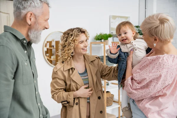 Весёлый ребёнок в джинсовой куртке машет рукой рядом с улыбающейся мамой, бабушкой и дедушкой в гостиной — стоковое фото