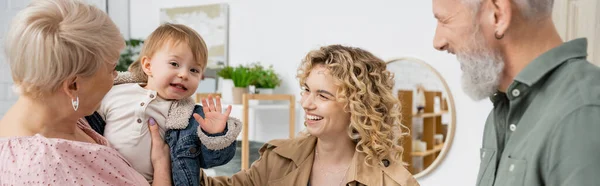 Беззаботная девушка машет рукой рядом с счастливыми бабушкой и дедушкой и улыбается матери в гостиной, баннер — стоковое фото