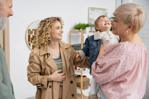Cheerful middle aged woman holding granddaughter in denim jacket near family in living room — Stock Photo