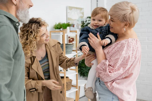 Щаслива блондинка в траншеї пальто посміхається біля дочки і батьків у вітальні — стокове фото