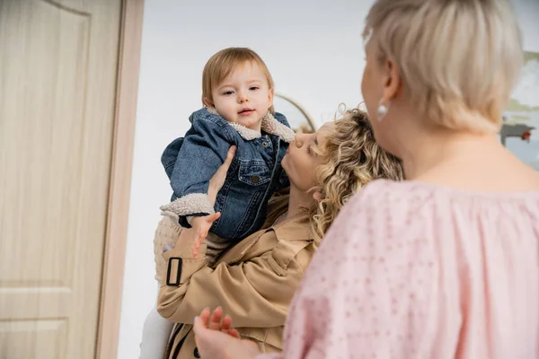 Blurred middle aged woman looking at daughter holding little girl in denim jacket near entrance door of apartment — Stock Photo