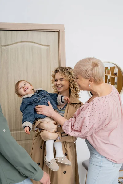 Mujer alegre sosteniendo hija pequeña en chaqueta de mezclilla cerca mientras visita a padres felices en casa - foto de stock