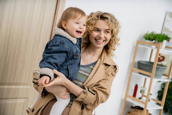Fröhliche blonde Frau mit welligem Haar, die ihre unbeschwerte Tochter in Jeansjacke in der Nähe der Haustür hält — Stockfoto
