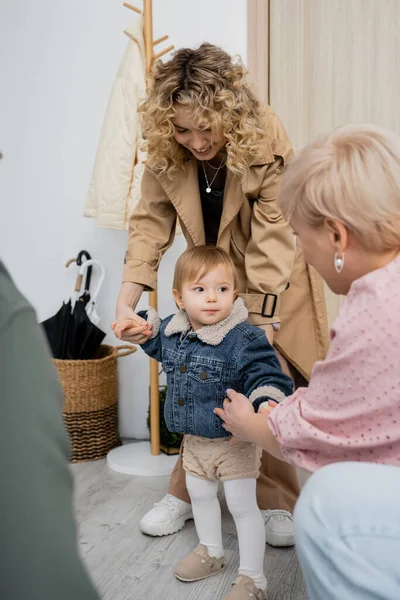 Femme souriante en trench coat et tout-petit fille en jean veste rendre visite aux grands-parents à la maison — Photo de stock