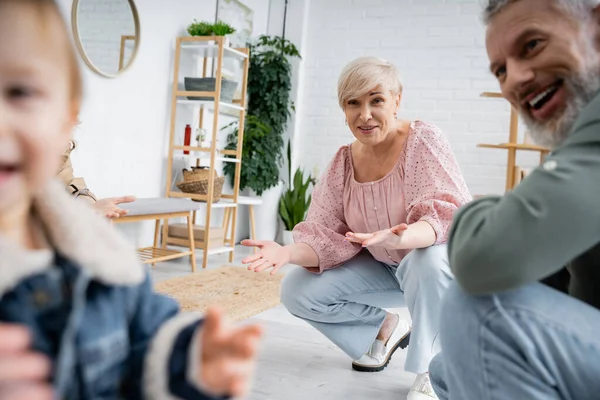 Overjoyed middle aged couple looking at blurred granddaughter in living room at home — Stock Photo