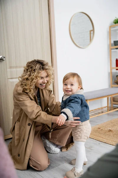 Curly woman in trench coat hugging carefree and cheerful daughter in denim jacket near door at home — Stock Photo