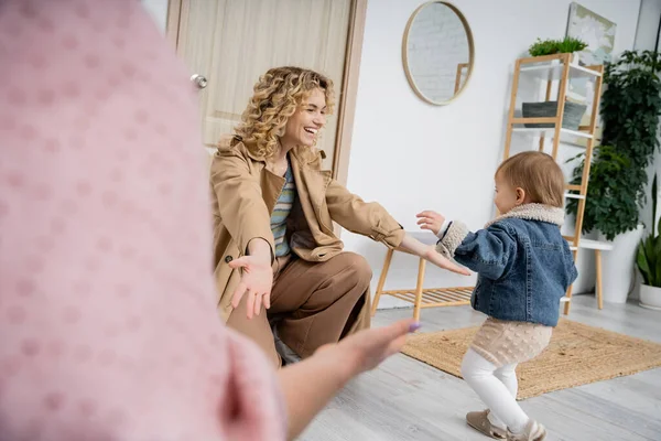Joyful woman in trench coat meeting daughter with outstretched hands near door at home — Stock Photo