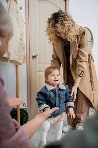 Blurred woman meeting daughter with overjoyed child in denim jacket near door at home — Stock Photo