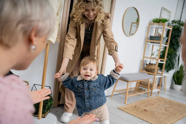 Счастливая женщина в плаще держит за руки малышку в джинсовой куртке во время визита к бабушке — стоковое фото