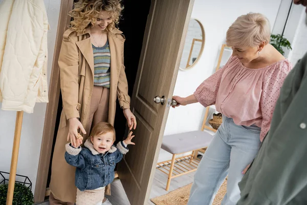 Feliz mujer de mediana edad abriendo la puerta a la hija con nieto en chaqueta de mezclilla durante su visita a casa - foto de stock