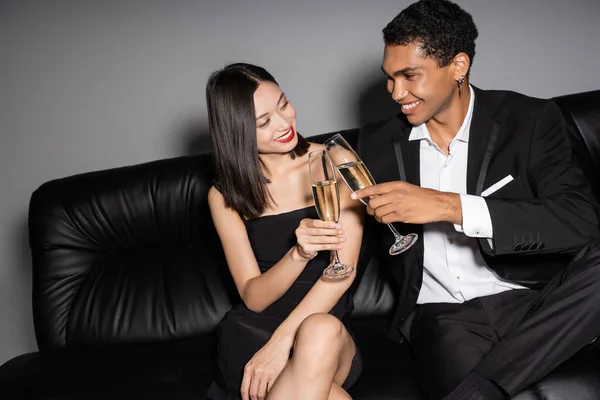Fröhliches und elegantes multiethnisches Paar auf schwarzer Ledercouch sitzend und Champagnergläser klirrend auf grauem Hintergrund — Stockfoto