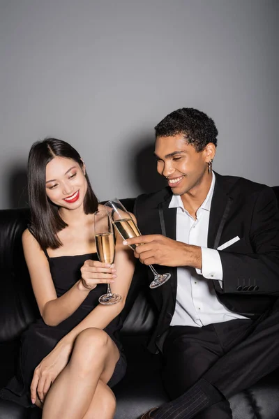 Счастливая межрасовая пара в черной элегантной одежде звон бокалов шампанского на кожаном диване на сером фоне — стоковое фото