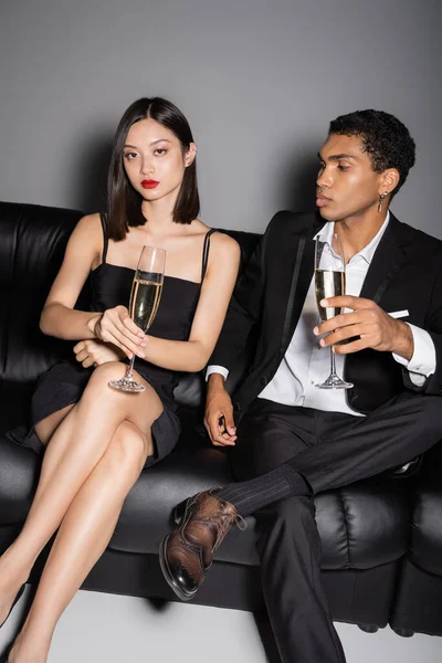 Elegant asian woman looking at camera near stylish african american man while sitting with champagne glasses on leather couch grey background — Stock Photo