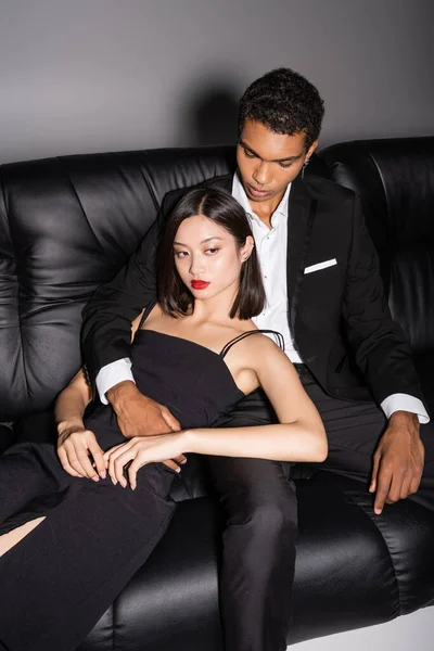 African american man in black suit embracing asian woman in strap dress on leather couch on grey background — Stock Photo