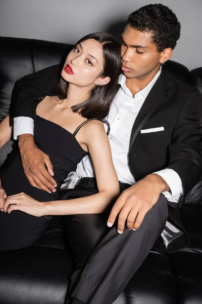 Sensual asian woman with red lips looking at camera near elegant african american guy on black couch on grey background — Stock Photo