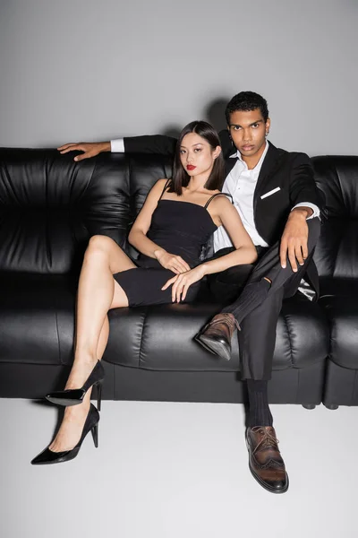 Comprimento total de mulher asiática em vestido de cinta e homem americano africano em terno preto olhando para a câmera no sofá de couro no fundo cinza — Fotografia de Stock