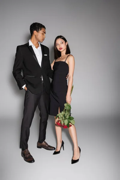 По всей длине африканский американец в черном стильном костюме обнимает азиатку с красными розами на сером фоне — стоковое фото