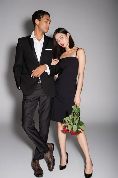 Comprimento total de mulher asiática com rosas vermelhas olhando para a câmera perto de homem americano africano elegante em fundo cinza — Fotografia de Stock