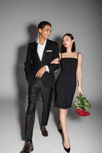 Longitud completa de hombre afroamericano feliz y elegante mujer asiática con rosas rojas caminando sobre fondo gris - foto de stock