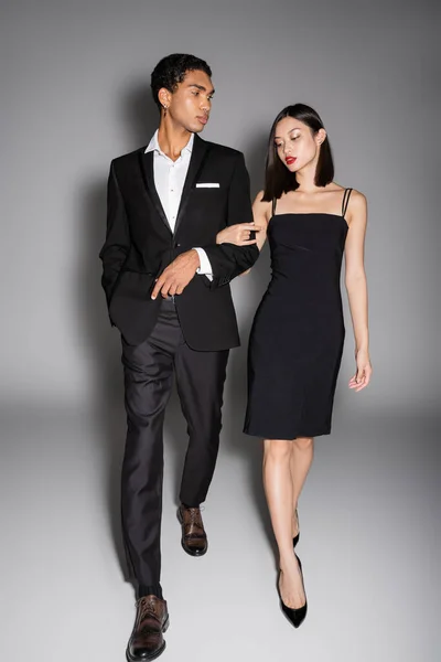 Longitud completa de la pareja multiétnica en traje elegante negro caminando sobre fondo gris - foto de stock