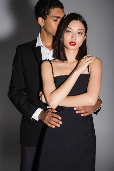 Joven africano americano hombre abrazando sensual asiático mujer en negro elegante vestido en gris fondo - foto de stock