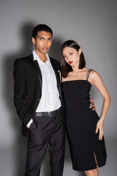 African american man in black suit and asian woman in strap dress looking at camera on grey background — Stock Photo