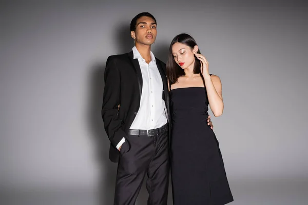 Africano americano hombre en elegante traje mirando cámara cerca asiático mujer en negro vestido en gris fondo - foto de stock
