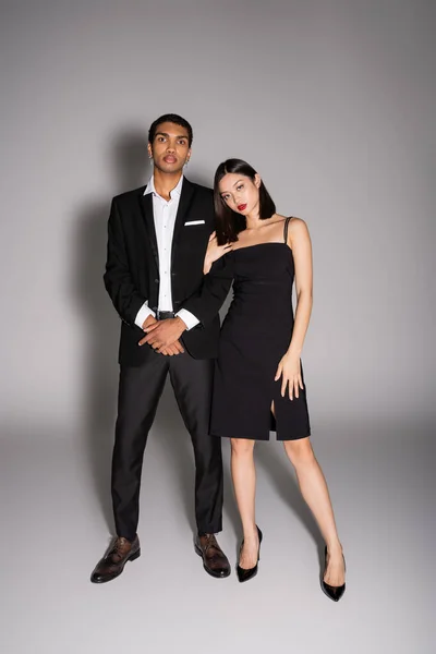 Longitud completa de la pareja interracial en elegante desgaste formal mirando a la cámara sobre fondo gris - foto de stock