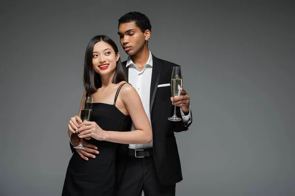Sonriente asiático mujer y elegante africano americano hombre holding champán copas aislado en gris - foto de stock