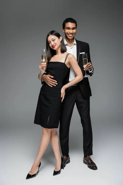 Piena lunghezza di allegro afro americano uomo abbracciando elegante donna asiatica mentre in piedi con bicchieri di champagne su sfondo grigio — Foto stock