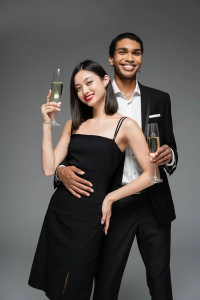Casal interracial em traje elegante segurando copos de champanhe e sorrindo para a câmera isolada em cinza — Fotografia de Stock