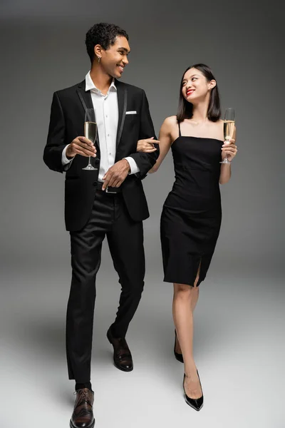 Full length of happy and elegant interracial couple walking with champagne glasses and looking at each other on grey background — Stock Photo