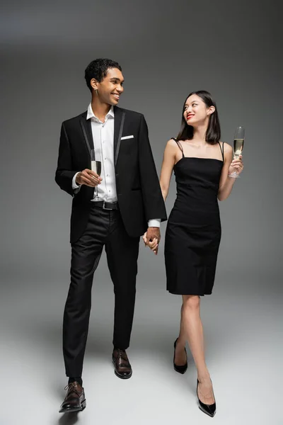 Comprimento total do elegante casal multiétnico com taças de champanhe de mãos dadas e sorrindo um para o outro enquanto caminham sobre fundo cinza — Fotografia de Stock
