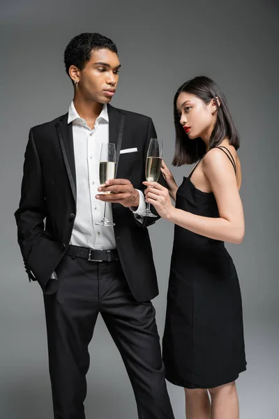 Élégant homme afro-américain en costume et femme asiatique en robe noire posant avec des verres de champagne isolé sur gris — Photo de stock