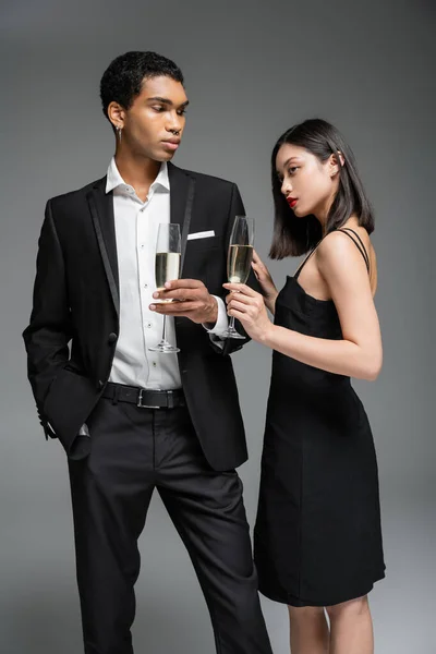 Elegante uomo africano americano in abito e donna asiatica in abito nero in posa con bicchieri di champagne isolati su grigio — Foto stock