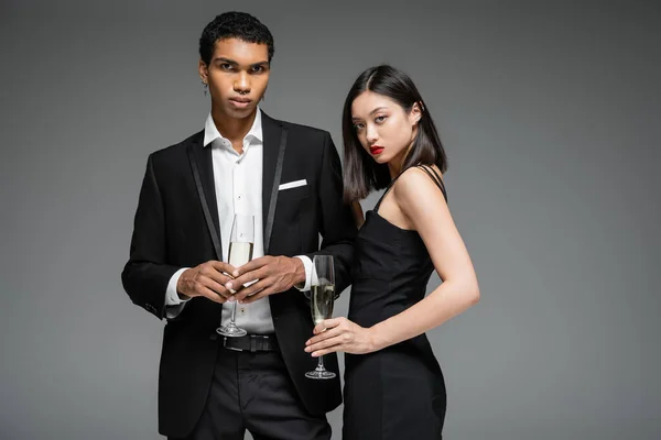 Africano americano uomo in nero vestito e asiatico donna in cinghia vestito tenere champagne e guardando fotocamera isolato su grigio — Foto stock