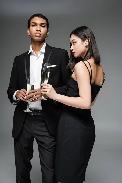 Giovane coppia interrazziale in abiti eleganti neri in posa con bicchieri di champagne isolati su grigio — Foto stock