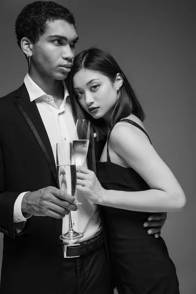 Pareja interracial joven y de moda en ropa formal sosteniendo copas de champán aisladas en gris - foto de stock