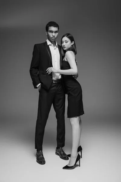 Longitud completa de hombre afroamericano en traje elegante y mujer asiática en vestido negro mirando a la cámara sobre fondo gris - foto de stock
