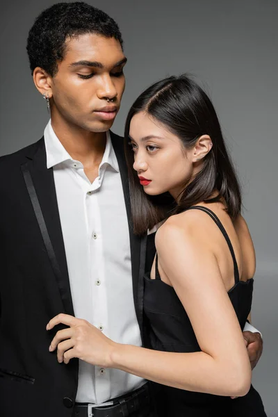 Elegant african american guy in black blazer embracing sensual asian woman looking away isolated on grey — Stock Photo